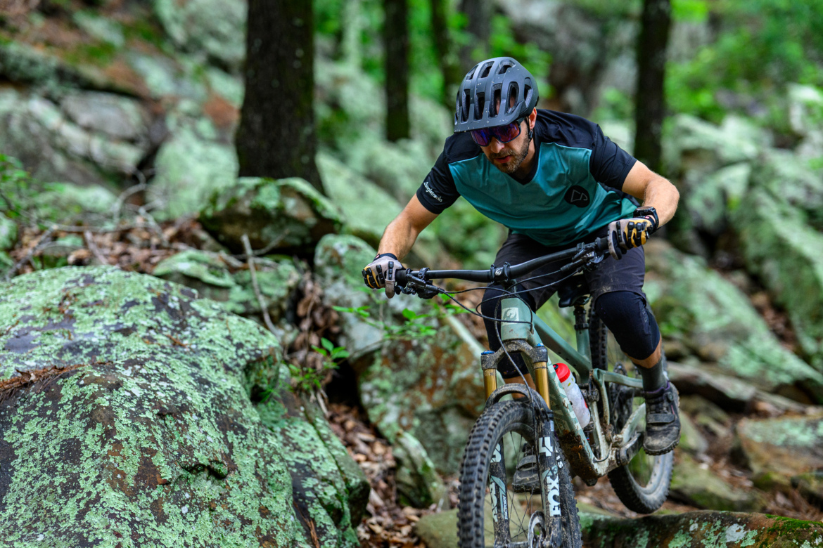 A mountain biker navigating rocky terrain in a wooded area, wearing a helmet and protective gear. The rider is focused and leaning forward on the handlebars as they maneuver over the uneven ground, surrounded by lush greenery and large, moss-covered rocks.
