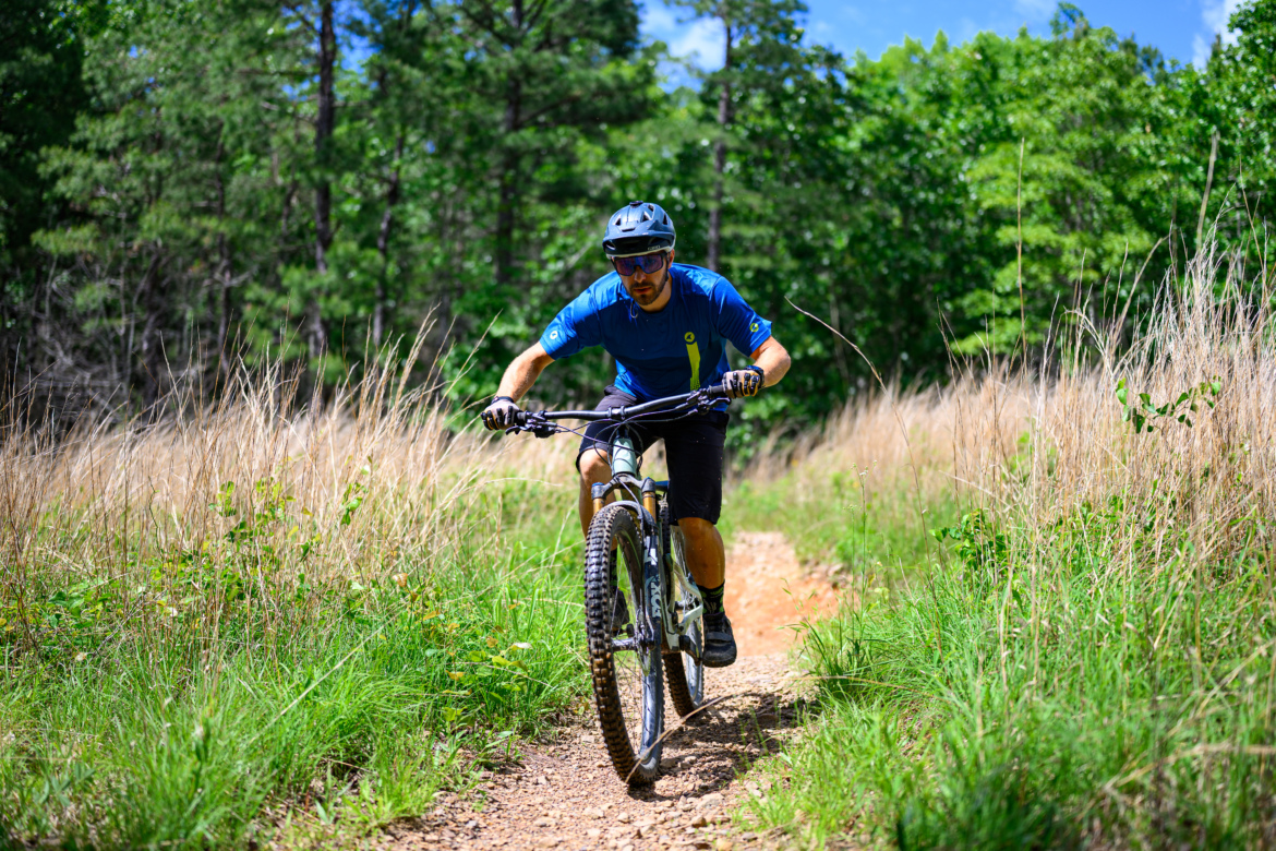 A mountain biker navigating a dirt trail surrounded by tall grass and trees on a sunny day. The rider is focused, wearing a blue jersey, helmet, and sunglasses, in an active riding position on a mountain bike.