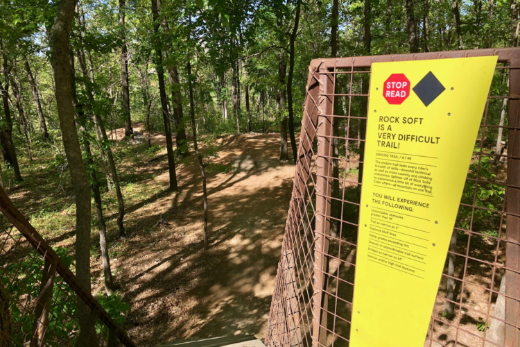 A close-up view of a warning sign at the entrance of a wooded trail, indicating that "Rock Soft" is a very difficult trail for biking with a list of potential obstacles and challenges. The background shows a winding dirt path through lush green trees.