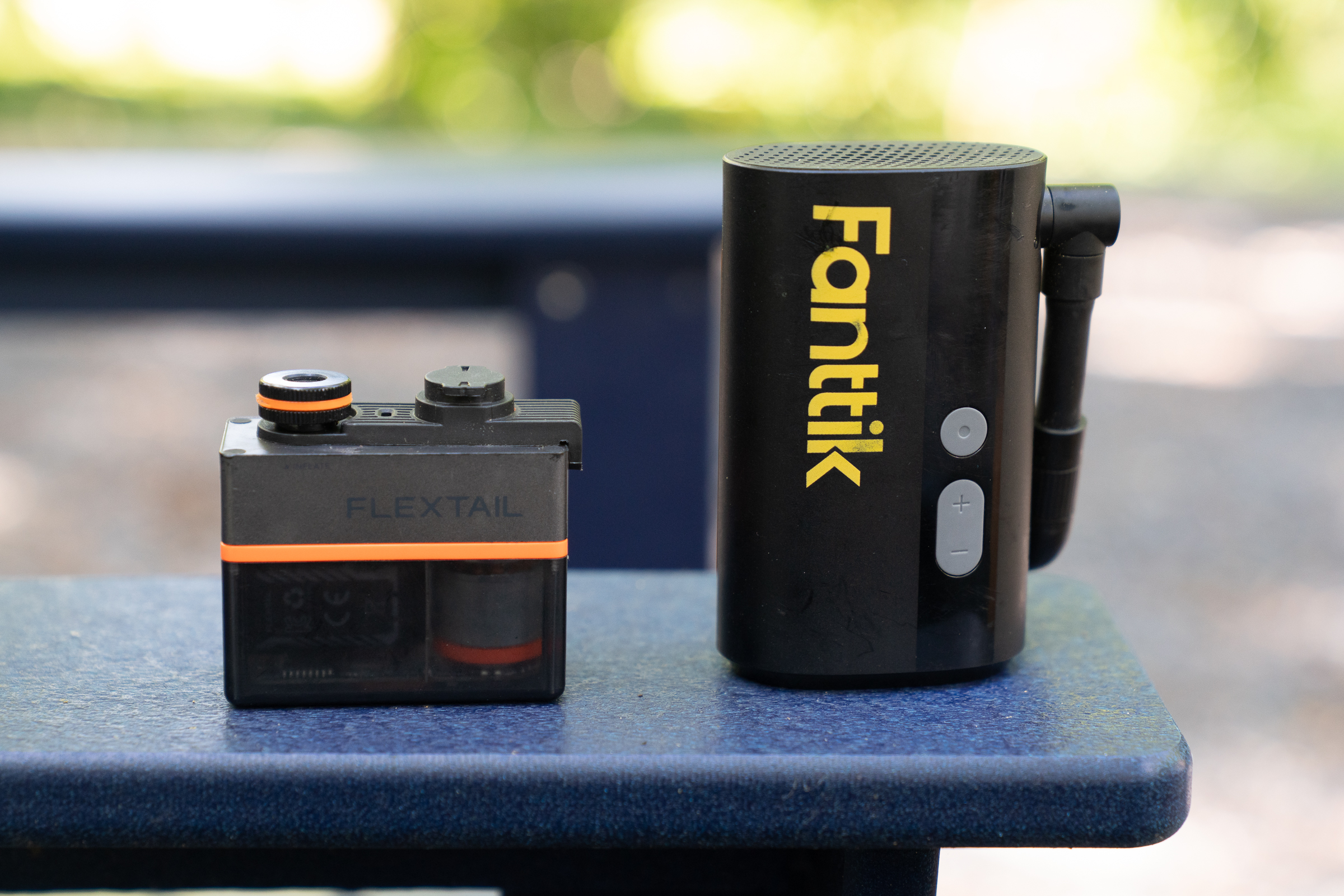 A small FLEXTAIL camera alongside a Fanttik device, both placed on a blue surface outdoors with a blurred natural background.