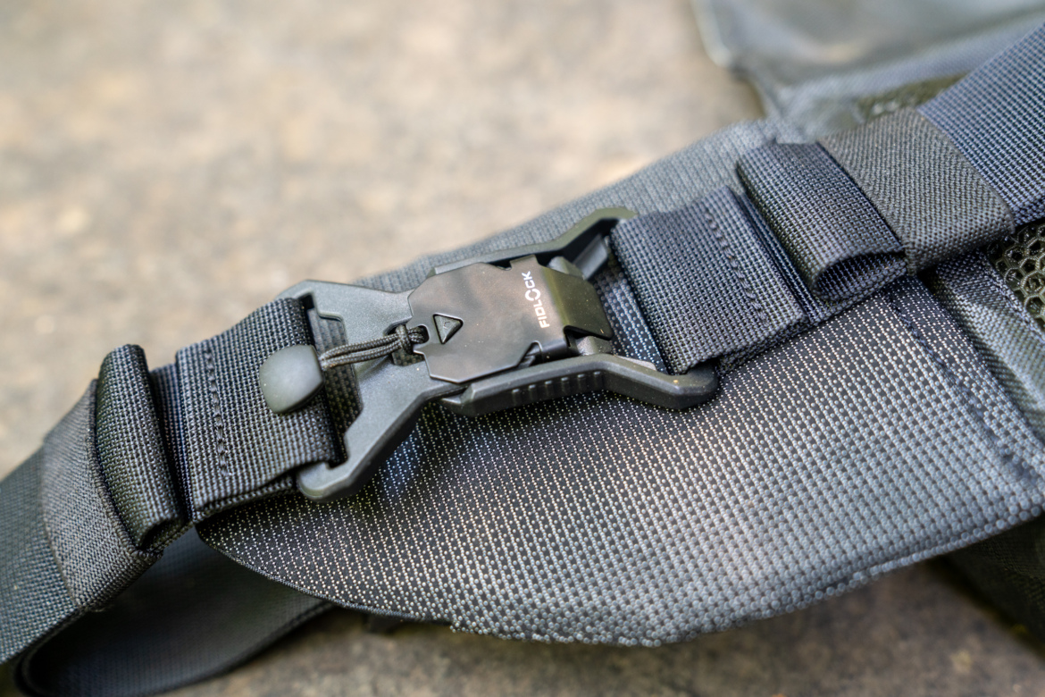 Close-up of a black adjustable buckle and strap, featuring a FIDLOCK clasp, resting on a textured surface. The strap showcases a durable fabric with a mesh section.