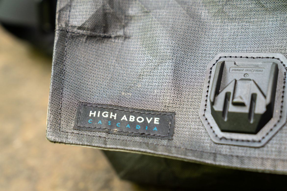 Close-up of a textured fabric featuring a label that reads "HIGH ABOVE CASCADIA," accompanied by a black plastic attachment on the side.