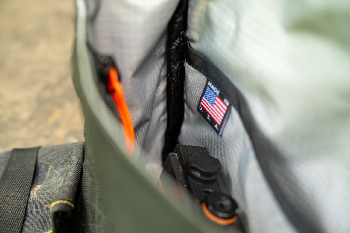 A close-up view of the interior of a bag, showing a label with the American flag and the text "MADE IN U.S.A." An orange cord is visible alongside dark gear or tools, set against a gray fabric lining. The background includes a blurred portion of the bag's exterior.