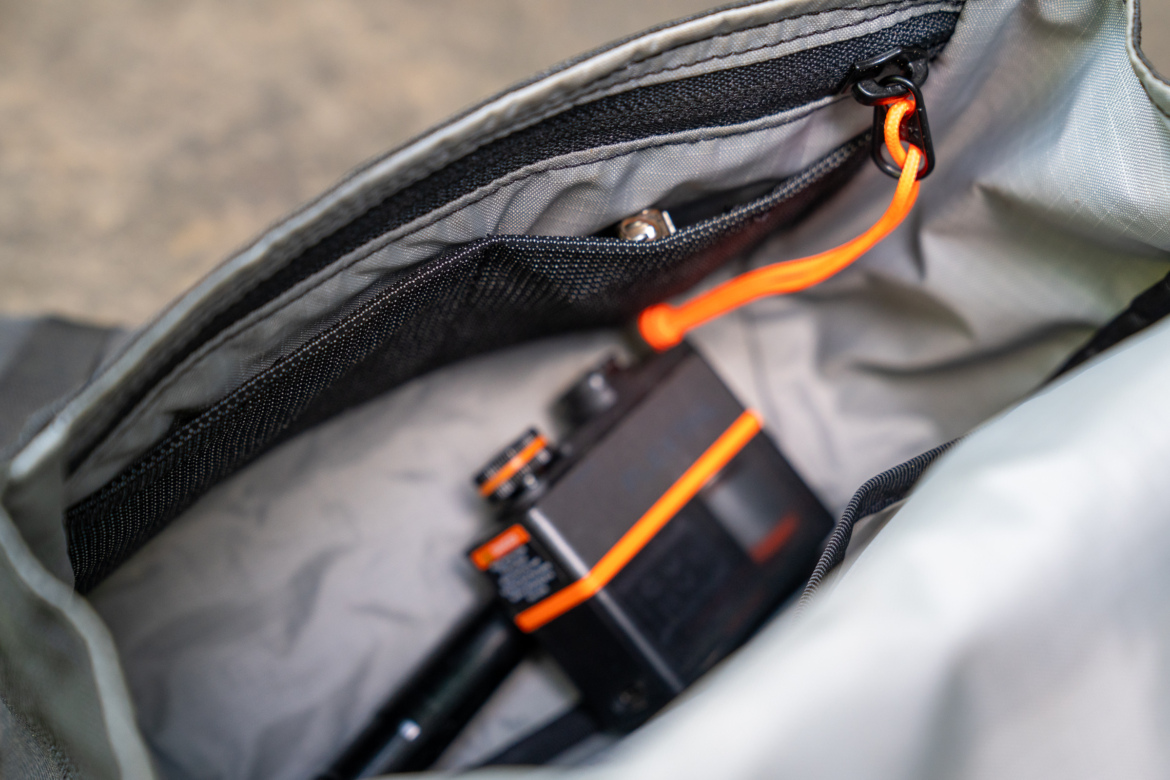 Close-up view of the interior of a bag, featuring a compact black camera partially visible with an orange accent. The bag has a gray lining and a zippered pocket with a bright orange cord.