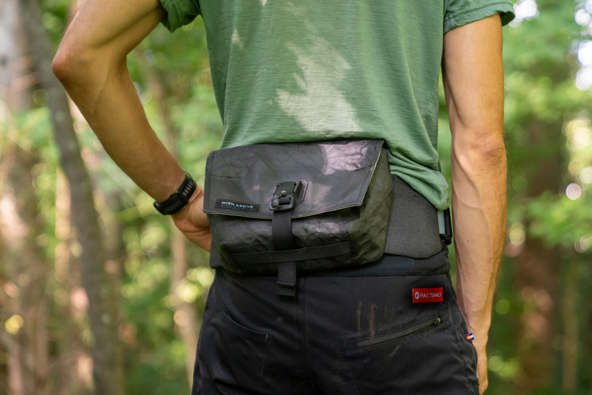 A person standing outdoors with their back to the camera, wearing a green shirt and dark pants. They have a black High Above Venture 3D hip pack secured at their waist with a buckle, featuring a label that reads "HIGH ABOVE." The background shows a blurred forest landscape with sunlight filtering through the trees.