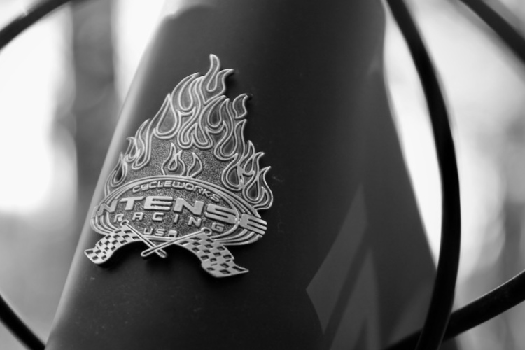 Close-up of a silver metal badge featuring a flame design and the text "Cycleworks Intense Racing USA," affixed to the frame of a bicycle. The image is in black and white, highlighting the details of the badge against a blurred background.