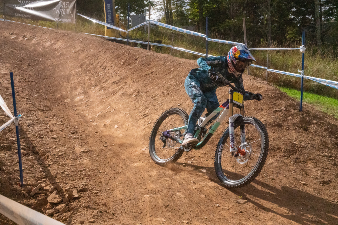 A mountain biker in blue and black gear navigates a dirt track, leaning into a corner as dust is kicked up from the ground. The scene captures the excitement and speed of downhill biking in a wooded area, with race banners in the background.