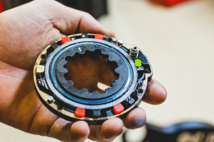 A close-up image of a mechanical component held in a hand, featuring a circular metal piece with a star-shaped opening in the center. The component has red and green accents and appears to be part of a larger mechanical assembly. The background is blurred, suggesting an industrial or workshop setting.
