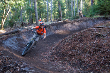 A mountain biker in an orange shirt and helmet navigates a dirt bike trail through a forest, leaning into a turn with a smile. The trail is surrounded by trees and fallen leaves, showcasing a vibrant outdoor setting. In the background, another rider can be seen. Demo Forest Flow Trail mountain bike trail.