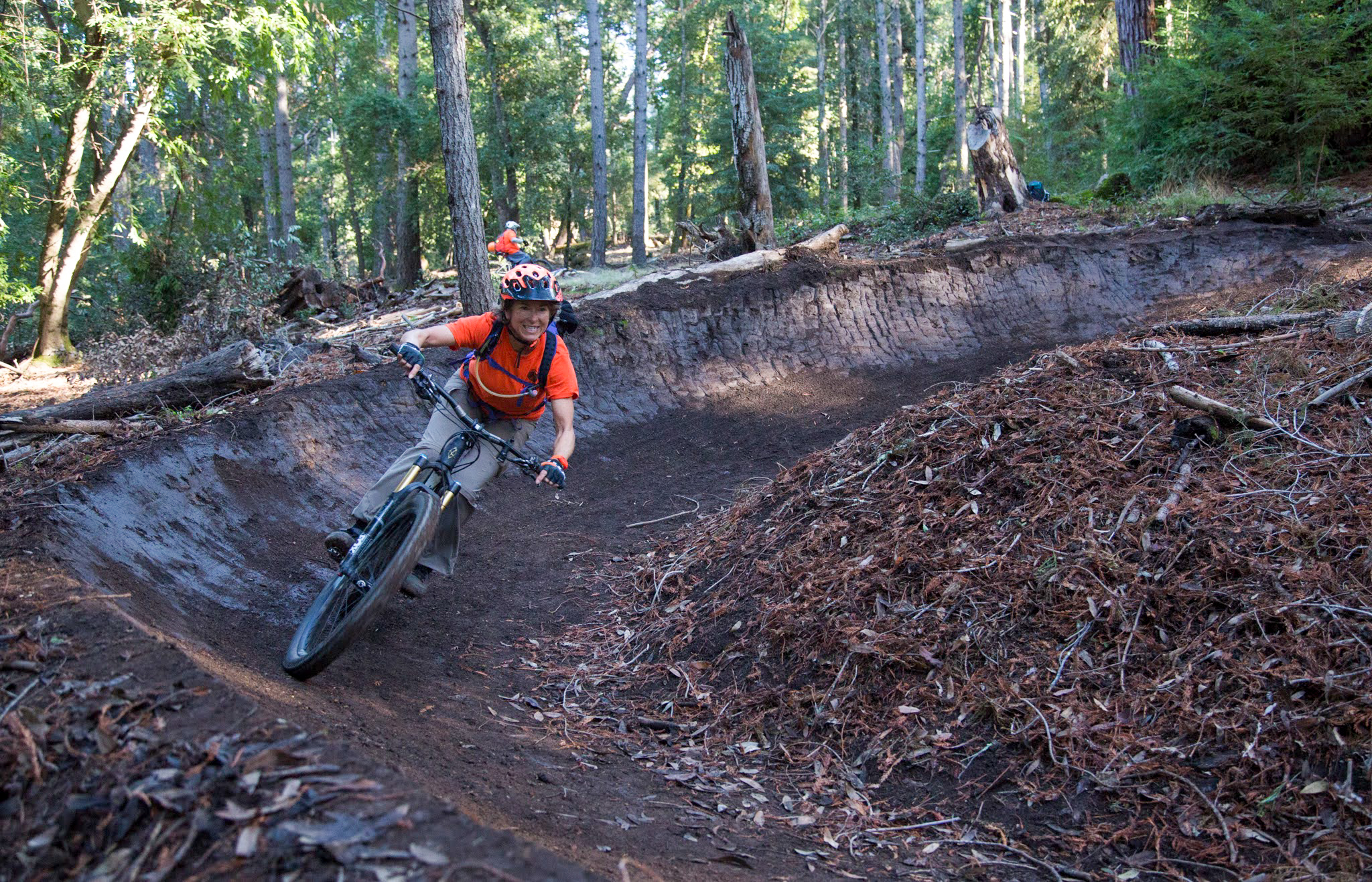 A mountain biker in an orange shirt and helmet navigates a dirt bike trail through a forest, leaning into a turn with a smile. The trail is surrounded by trees and fallen leaves, showcasing a vibrant outdoor setting. In the background, another rider can be seen. Demo Forest Flow Trail mountain bike trail.