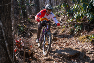 A mountain biker with sunglasses and a colorful jersey navigates a rocky trail in a wooded area, focused on maintaining balance as he rides downhill. The ground is covered with leaves and scattered rocks, while trees and greenery surround the path. Farlow Gap mountain bike trail.