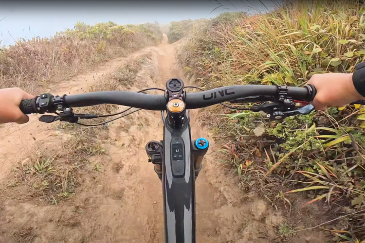 A view from the handlebars of a mountain bike navigating a dirt trail surrounded by tall grass and vegetation. The bike displays a digital dashboard, and the terrain appears rocky and uneven, suggesting an adventurous ride.