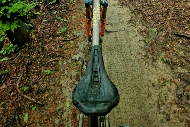 Alt text: A top-down view of a mountain bike saddle with a muddy trail and foliage visible in the background, indicating a ride through a natural setting.
