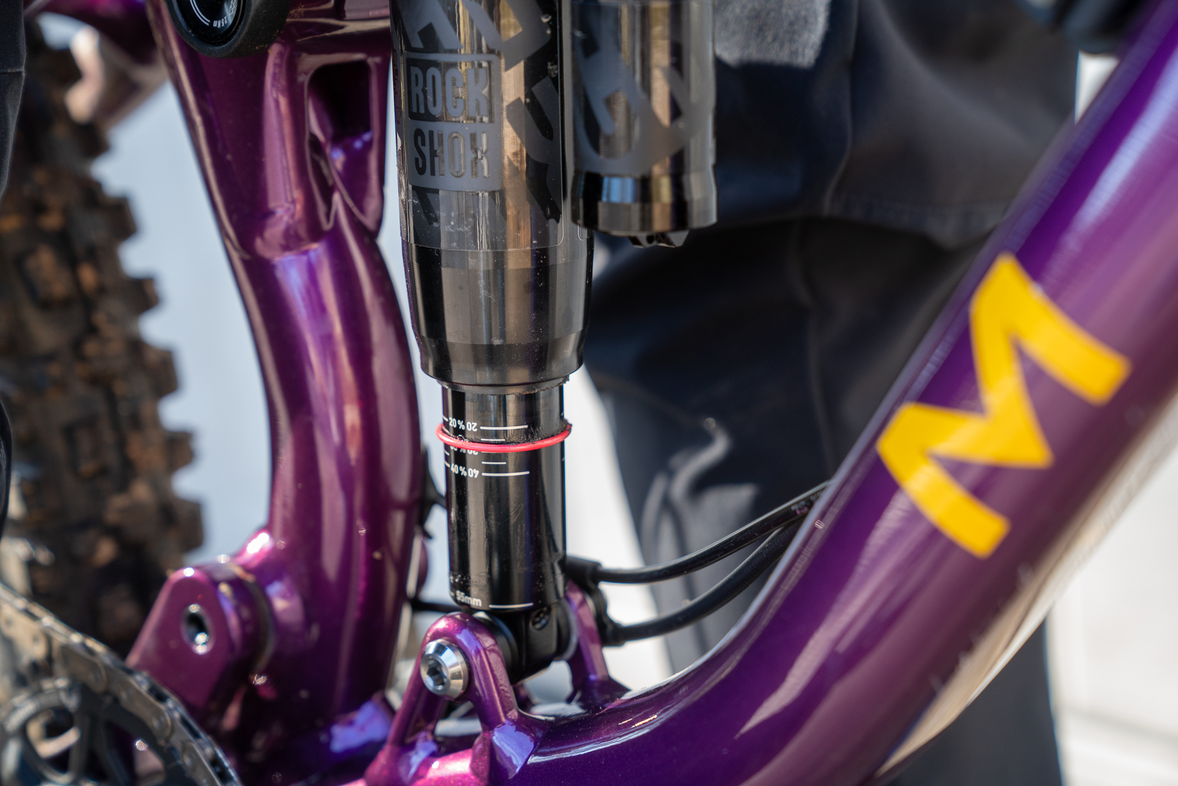Close-up of a purple mountain bike frame featuring a RockShox suspension system. The image highlights the shock absorber with adjustment markings and a prominent red line. The bike's frame design includes a bold yellow "M" logo, with a glimpse of the rear wheel's tread in the background.