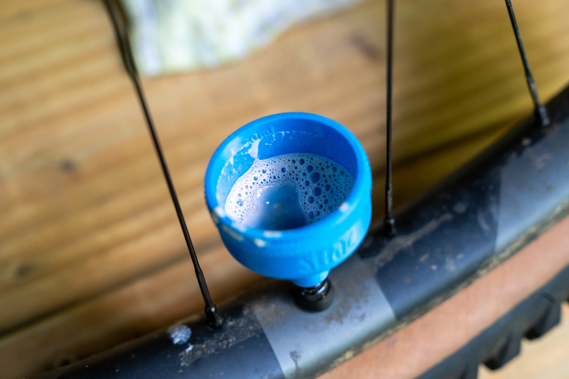 A close-up image of a blue valve cap sitting on a bicycle wheel, filled with soapy water, with part of the wheel visible in the foreground and a wooden surface in the background.