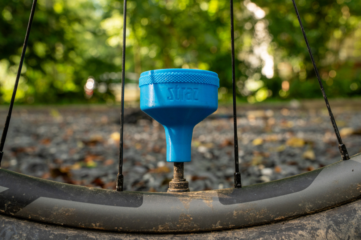 A close-up view of a blue valve cap attached to a bicycle wheel, surrounded by black spokes. The background features blurred greenery and gravel, indicating an outdoor setting. The valve cap is prominently displayed, showcasing its texture and branding.