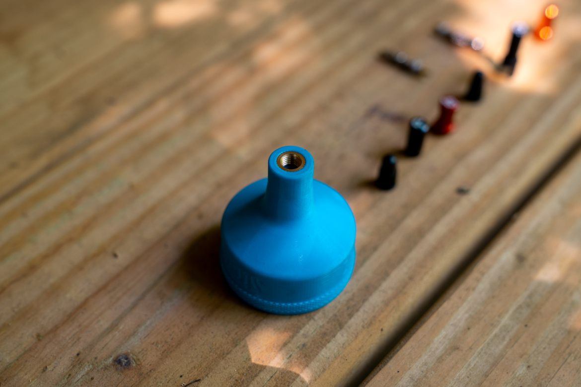 A blue funnel with a threaded opening sits on a wooden surface, with several colored caps (black, red, and orange) arranged in a line behind it.