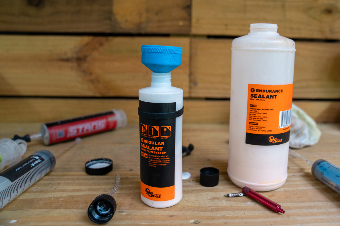 An assortment of sealant containers and tools on a wooden surface. The foreground features a bottle labeled "Regular Sealant" with a blue cap and a bottle labeled "Endurance Sealant." Various syringes and a red tool are scattered around them, alongside a black cap and a cloth. The background consists of wooden panels, adding a rustic feel to the workspace.
