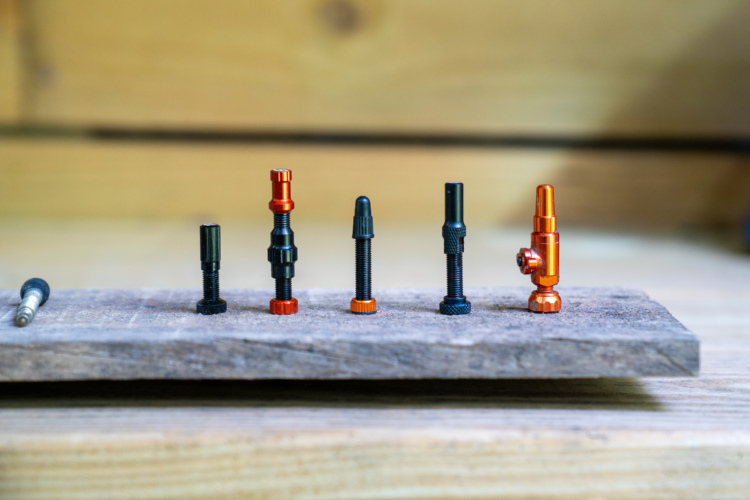 A close-up image of a wooden plank displaying a row of various metal and anodized components, including black and orange parts in different shapes and sizes. The items consist of screws, bolts, and fittings, highlighting their intricate designs and finishes against a blurred wooden background.