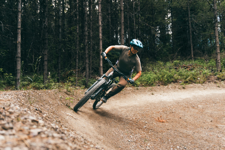 A person riding a mountain bike on a dirt trail, leaning into a turn with trees in the background. The individual is wearing a helmet and protective gear, showcasing an active outdoor biking scene.