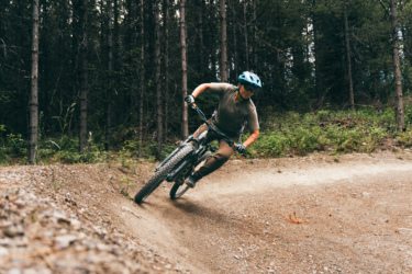 A mountain biker leaning into a turn on a dirt trail surrounded by tall trees. The rider is wearing a helmet and biking gear, with one foot raised off the ground as they navigate the curve of the path. Mount Harry Davis mountain bike trail.