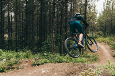 A mountain biker performing a jump on a dirt trail surrounded by tall pine trees and greenery. The cyclist, wearing a helmet and athletic attire, is airborne above the trail, showcasing a dynamic action shot in a forested setting. Mount Harry Davis mountain bike trail.