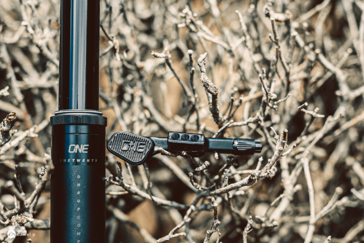 A close-up view of a black bike dropper seat post with an attached remote lever, set against a background of dry, tangled branches. The seat post features the brand name "ONE" along with the model designation "TWENTY."