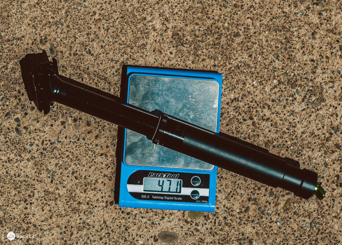 A black bicycle dropper post resting on a digital scale, displaying a weight of 477 grams, against a textured concrete surface.