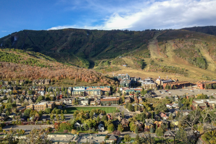 A panoramic view of a mountain town nestled at the foot of a hillside, featuring a mix of residential houses and commercial buildings. Vibrant fall foliage decorates the landscape, and ski slopes can be seen in the distance, under a clear blue sky with wispy clouds.