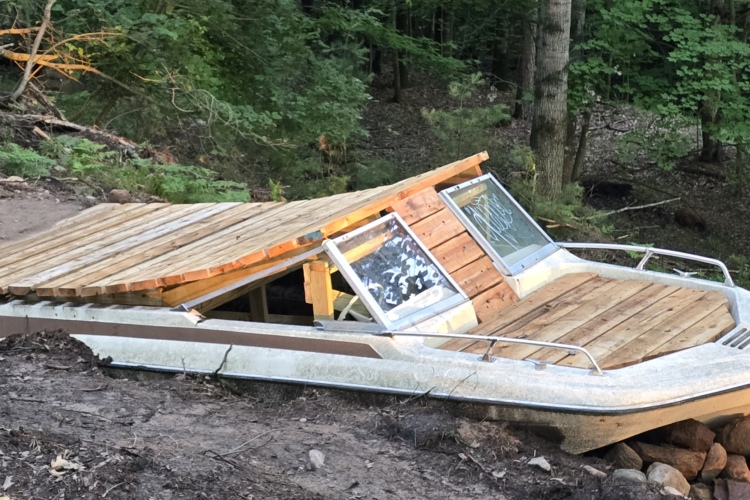 A partially submerged boat on a muddy shoreline, featuring a wooden deck on top and positioned among trees in a forested area. The boat's windows are visible, indicating its former use, while the surrounding environment is lush and green.