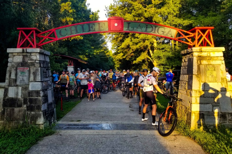 A large group of cyclists gathers at the entrance to the Chief Ladiga Trail, marked by a prominent red archway. The scene is set in a wooded area with lush green trees in the background, and the sun casts a warm glow on the participants. Some cyclists are preparing their bikes while others converse, creating a lively atmosphere in anticipation of a ride.