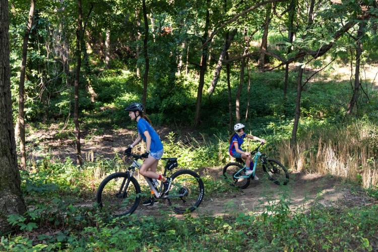 Two children riding mountain bikes on a dirt trail in a lush green forest. The older child, wearing a blue t-shirt and helmet, is in the foreground, while the younger child, dressed in a colorful cycling outfit and helmet, follows behind. Sunlight filters through the trees, creating a vibrant outdoor scene.