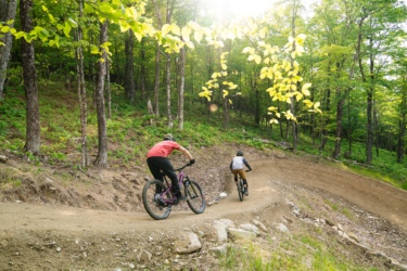 Two mountain bikers navigating a winding dirt trail in a lush, green forest under sunlight, surrounded by trees and foliage. Sugarloaf Bike Park mountain bike trail.