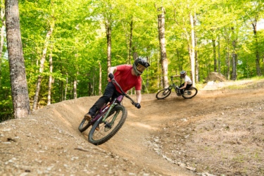 Two mountain bikers navigate a dirt trail in a wooded area. One rider is leaning into a turn, wearing a red shirt and helmet, while the other rides closely behind in a white shirt. The scene is surrounded by lush green trees, and the trail is well-defined with packed dirt and gravel. Sugarloaf Bike Park mountain bike trail.