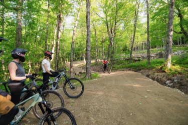 Three mountain bikers stand by their bikes on a dirt trail surrounded by lush green trees. One rider is in motion, navigating a curve on the trail. The scene captures the essence of outdoor biking in a forested area. Sugarloaf Bike Park mountain bike trail.