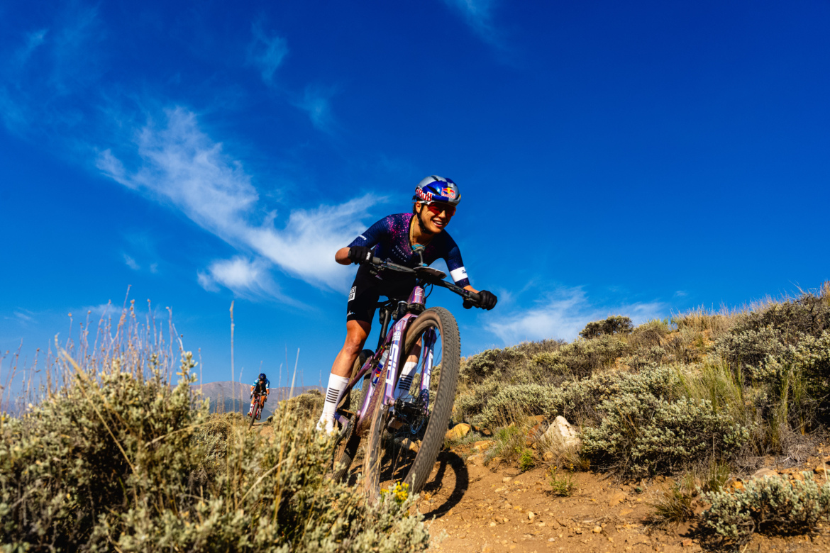 A mountain biker rides on a dirt trail surrounded by sparse vegetation, displaying a joyful expression and wearing a helmet and colorful cycling gear. The bright blue sky adorned with wispy clouds creates a vibrant backdrop. Another cyclist can be seen in the background on a similar path.