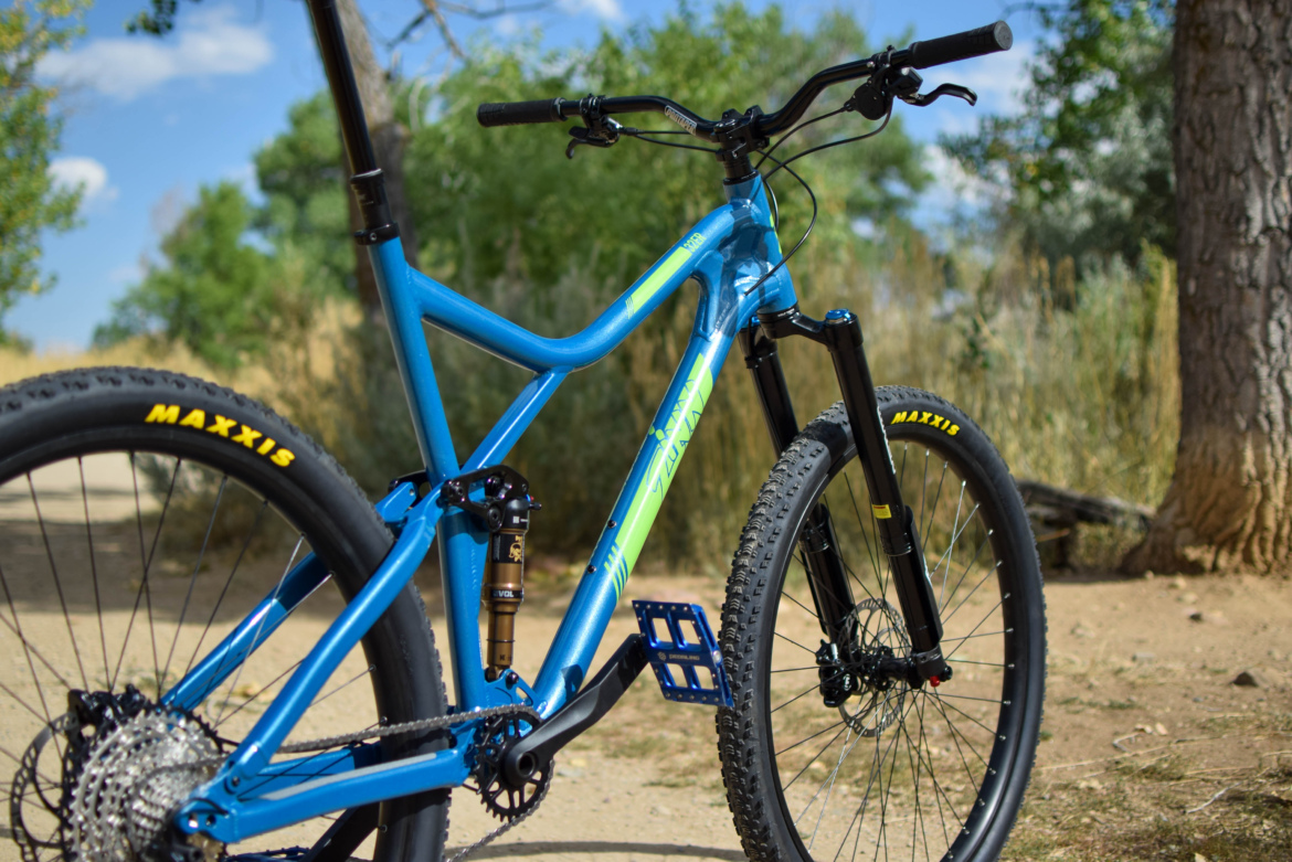 Blue mountain bike with green accents parked on a dirt trail, surrounded by trees and vegetation under a clear sky. The bike features thick tires and a modern design, showcasing detailed components like the suspension and pedals.
