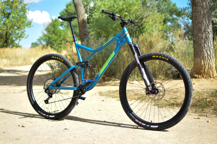 A blue mountain bike with green accents, positioned on a dirt path surrounded by trees and tall grass, under a clear blue sky. The bike features thick tires, disc brakes, and a suspension fork, perfect for trail riding.