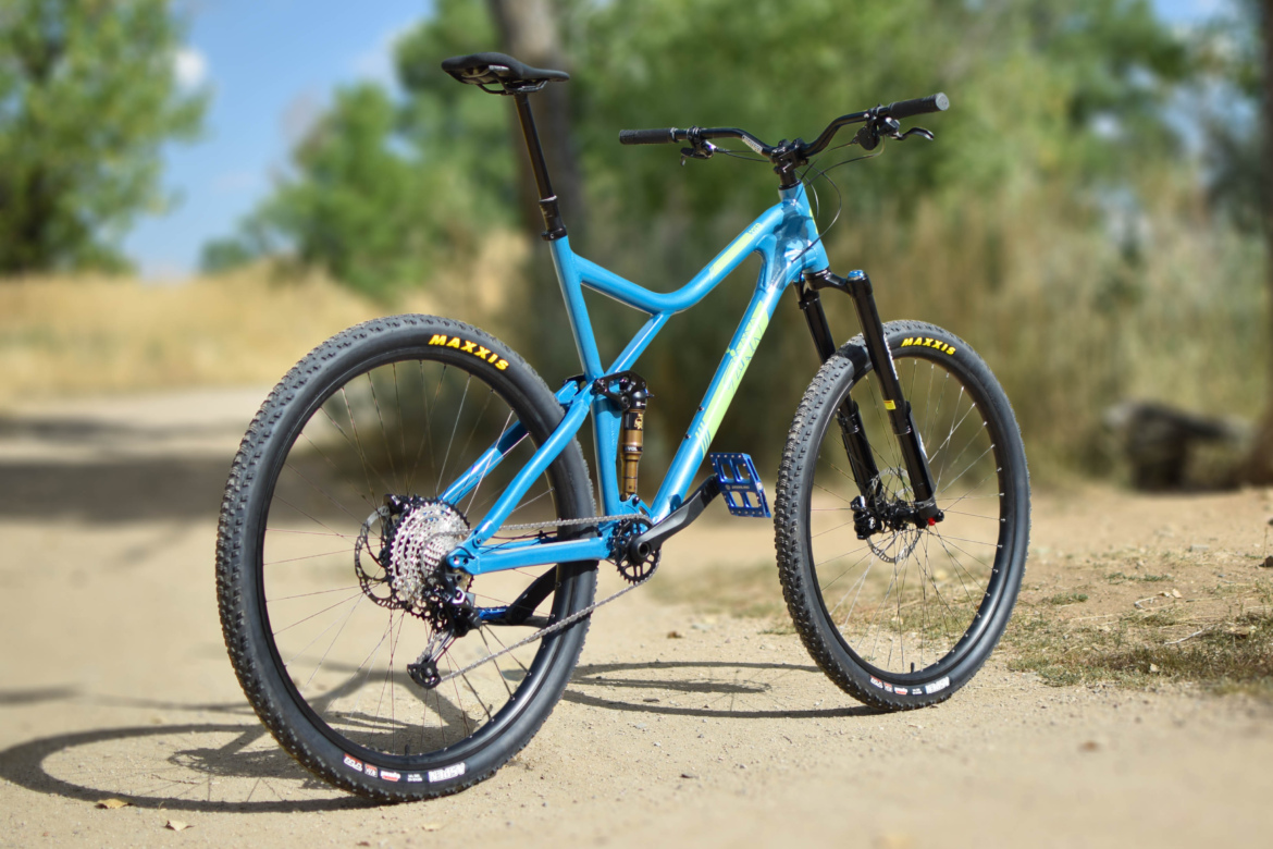 A blue 32er full suspension mountain bike with black wheels and yellow Maxxis tires is parked on a dirt path surrounded by green foliage. The bike features a suspension system, a sleek frame design, and is positioned slightly angled to showcase its details. Soft sunlight filters through the trees in the background.