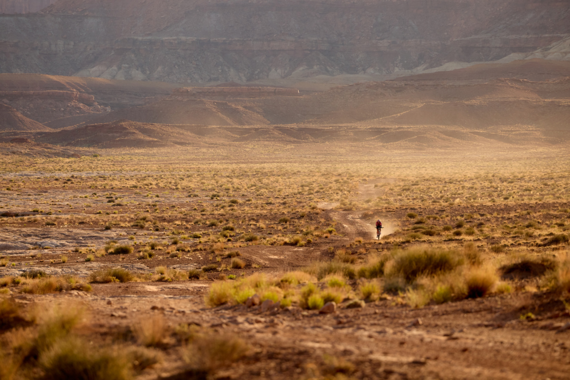 A lone figure walking along a dusty path in a vast, arid landscape with rolling hills and scattered vegetation, illuminated by soft golden light during sunset or sunrise.