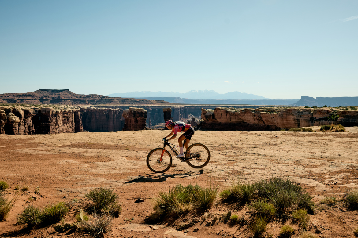 A mountain biker riding on a rocky landscape with rugged cliffs in the background under a clear blue sky. The terrain features a mix of sandy earth and sparse vegetation, showcasing a beautiful outdoor setting for biking.