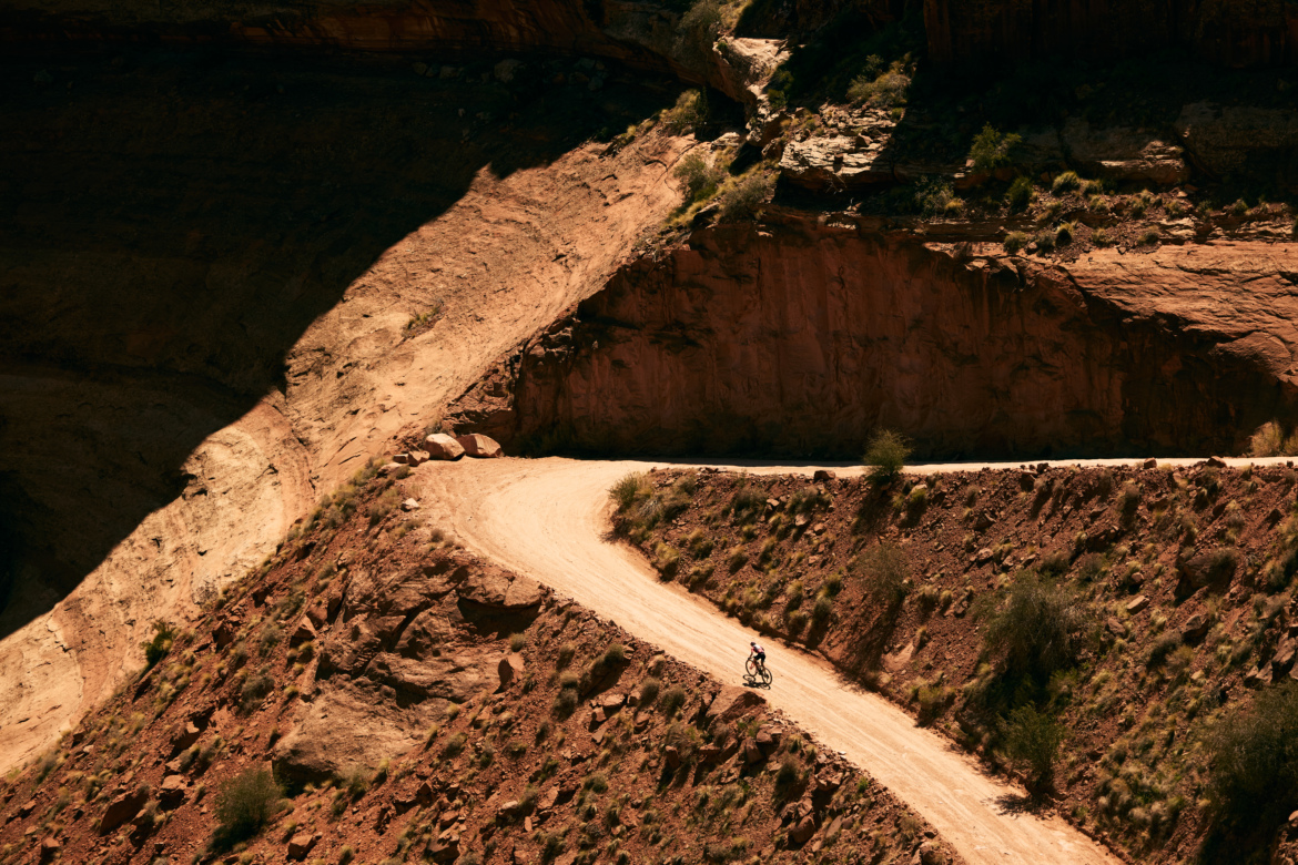 A cyclist riding on a winding dirt trail through a rugged, reddish-brown landscape surrounded by steep cliffs and shadows.