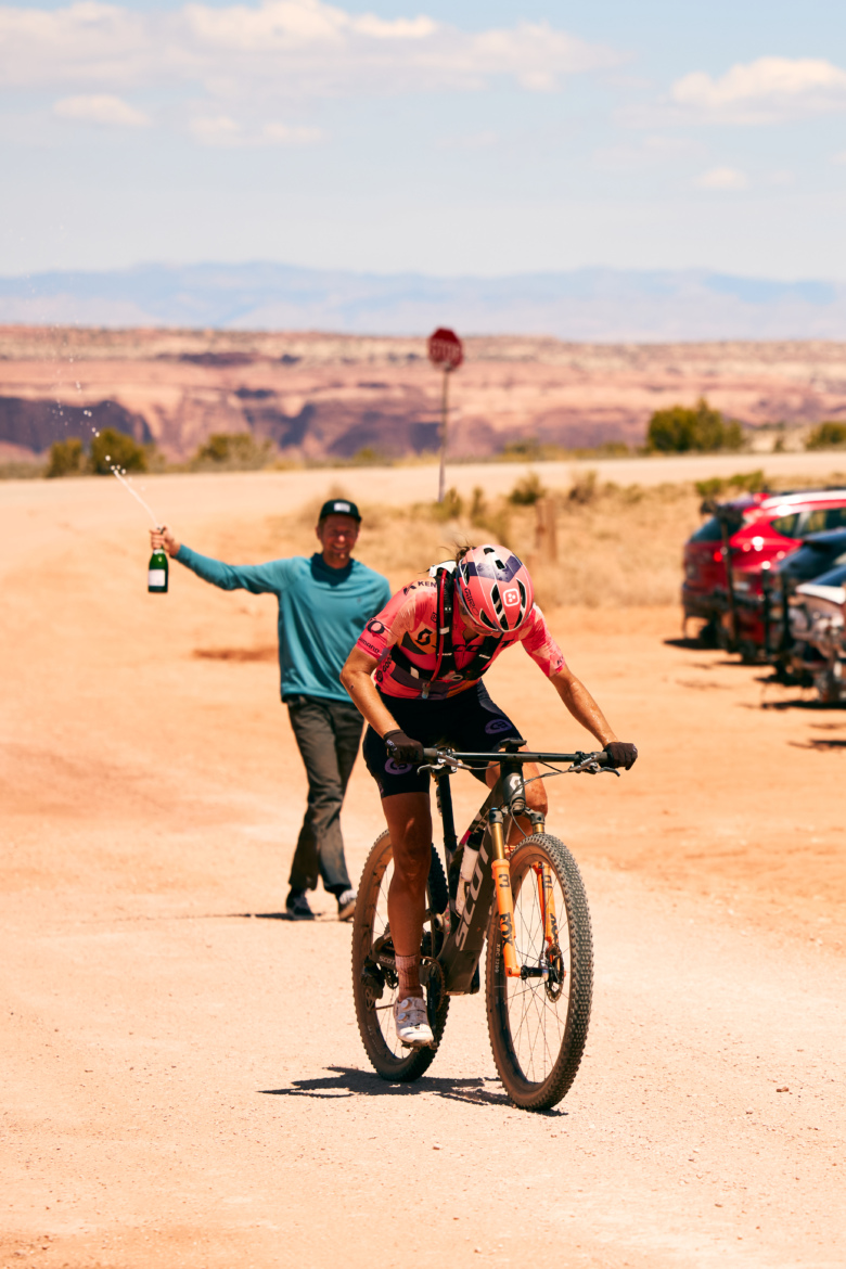 A mountain biker wearing a pink jersey rides on a dirt road, while a person in a blue shirt and cap stands in the background, celebrating by spraying champagne. Cars are parked nearby, and a scenic landscape is visible in the distance under a blue sky.