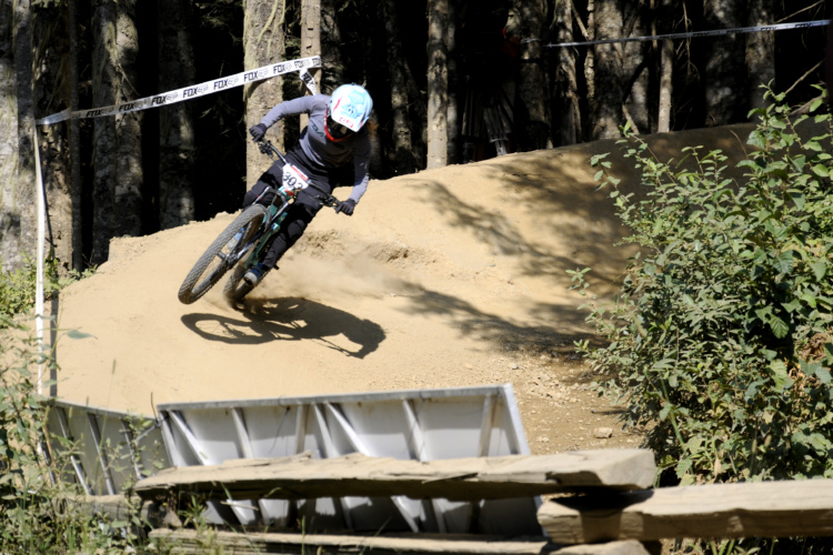 A mountain biker navigates a dirt track while leaning into a turn, with dust kicked up from the tires. Surrounding trees and a wooden structure are visible in the background.