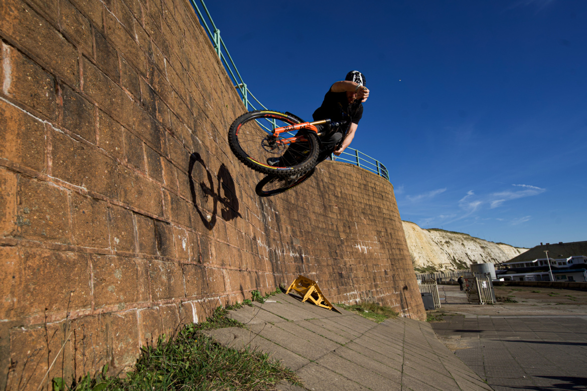 A mountain biker performs a wall ride against a textured stone wall, captured mid-air with one wheel off the ground. The scene is set against a clear blue sky, with the biker's shadow cast on the wall. In the foreground, there is an inclined pathway and some greenery, as well as a yellow safety barrier.