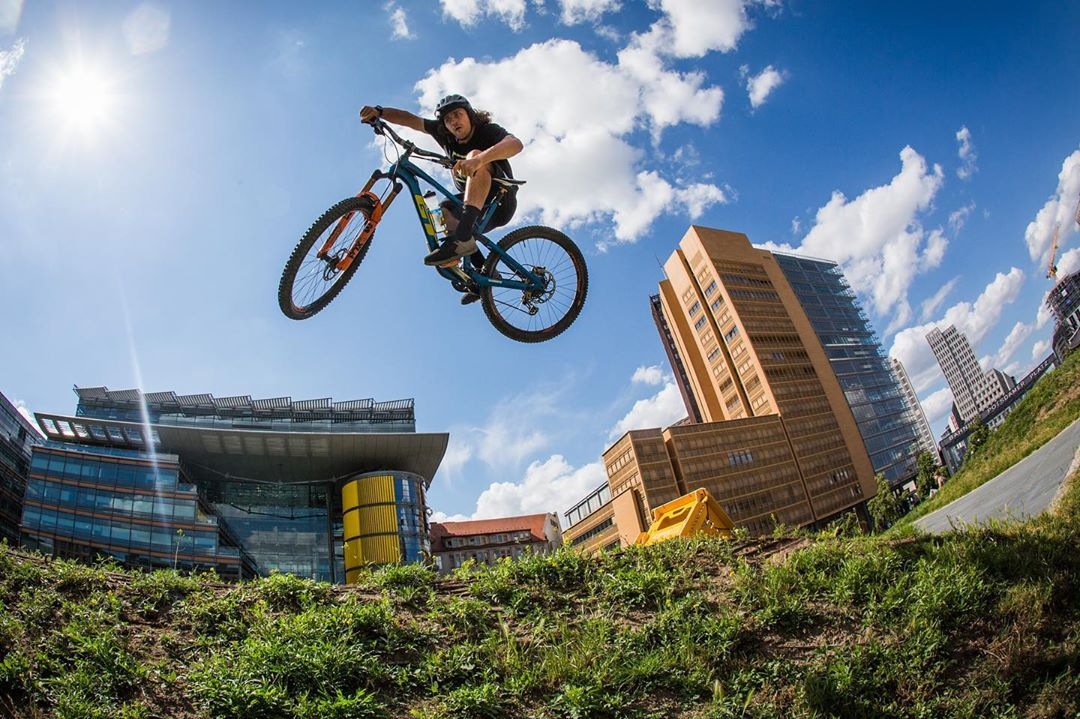 A mountain biker performs a jump in mid-air against a backdrop of urban buildings and a clear blue sky, with the sun shining brightly overhead.