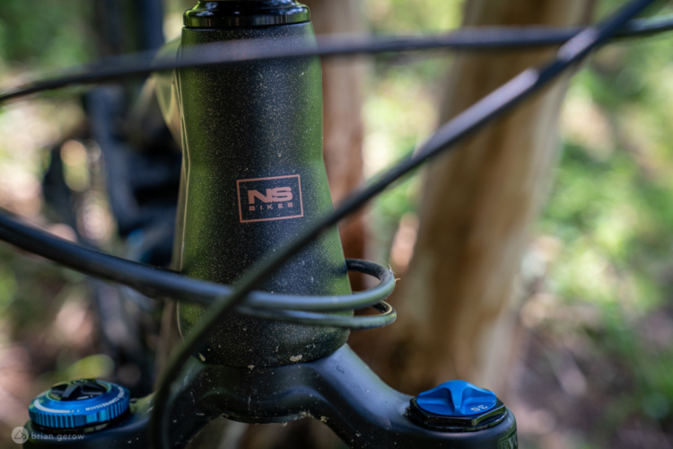 Close-up of a mountain bike's head tube featuring the "NS Bikes" logo, with visible cables and a blue adjustment knob on the fork. The background includes greenery and blurred tree trunks.