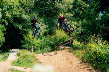 Two mountain bikers performing tricks in mid-air over a dirt jump, surrounded by lush greenery. The first rider, wearing a black helmet and dark clothing, is airborne on a teal bike, while the second rider in a yellow helmet is also airborne, showcasing dynamic action under a bright blue sky. A wooden ramp is visible in the background, enhancing the outdoor biking scene. DTE Energy Foundation Trails mountain bike trail.
