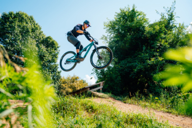 A mountain biker in athletic gear performs a jump over a dirt ramp, surrounded by lush greenery under a clear blue sky. DTE Energy Foundation Trails mountain bike trail.