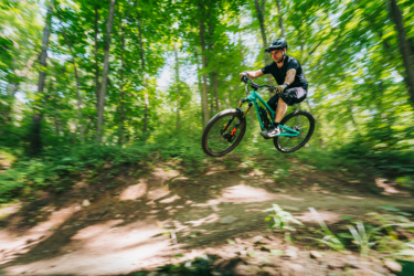 A mountain biker in mid-air jumps off a dirt ramp in a lush green forest, showcasing dynamic motion and excitement. The scene is filled with vibrant green foliage, emphasizing the outdoor environment. DTE Energy Foundation Trails mountain bike trail.